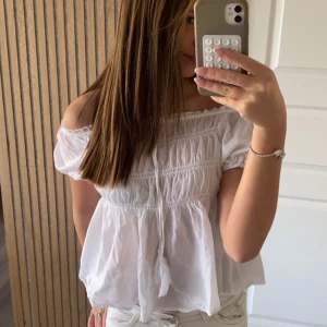 Vit offshoulder blus med knyt - Säljer en luftig vit blus med offshoulder och korta ärmar. Toppen har smockdetaljer över bröstet och ett sött knyt med tofs framtill. Den är tillverkad i tunn bomull och har en flowy passform som ger en chill vibe.