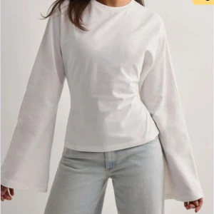 Vit omlottopp med vida ärmar - Supersnygg vit topp med vida långa ärmar och öppen rygg som knyts med en stor rosett baktill. Toppen har en croppad passform och är gjord i mjuk bomull. Perfekt för dig som vill ha en trendig och unik look.