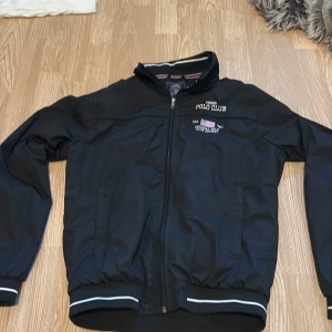 Svart vindjacka från Vinson Polo Club - Svart vindjacka från Vinson Polo Club med broderad logga och amerikansk flagga på bröstet. Jackan har dragkedja framtill, ribbade muddar med vit rand och ståkrage. Tillverkad i polyester och har en clean, sportig look.