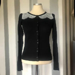 Svart kofta med spetskrage och knappar - Svart kofta med lång ärm och snygg vit spetsdetalj vid kragen. Tröjan har knappar hela vägen framtill och en slim passform som sitter snyggt på kroppen. Perfekt för dig som gillar klassisk stil med en twist.