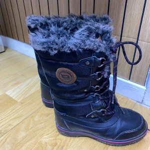 Svarta vinterstövlar från Leaf - Svarta vinterstövlar från Leaf med högt skaft, snörning och mysig grå fuskpäls runt kanten. Stövlarna har detaljer i skinn och textil samt en dekorativ rand i rosa på sulan. Perfekta för kalla dagar när du vill hålla stilen och värmen.