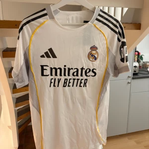 Real Madrid Mbappé fotbollströja - Snygg vit Real Madrid fotbollströja från Adidas med Mbappé och nummer 9 på ryggen. Tröjan har svarta detaljer, gula linjer längs sidorna och klubbmärke på bröstet. Materialet är lätt och andas, perfekt för match eller träning. Klassisk kortärmad modell med rund hals.