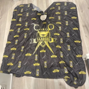 Svart barberarkappa med gul print - Barberarkappa i svart med coolt gult tryck och motiv av saxar, rakknivar och mustascher. Elastisk halsöppning för enkel användning. Perfekt för hemmaklippning eller barbershop-vibe. Materialet är lätt och syntetiskt, skyddar kläderna under klippning.