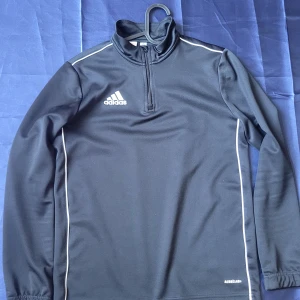 Svart Adidas långärmad träningströja - Svart långärmad träningströja från Adidas med vit logga på bröstet och vita detaljer längs ärmarna. Tröjan har en hög krage och half zip framtill. Materialet är polyester och modellen är slim fit, perfekt för sport och aktivitet.