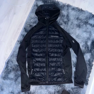 Svart cardigan Peak Performance S - Svart pufferjacka från Peak Performance med huva och dragkedja. Jackan har quiltade partier framtill och på huvan, samt släta ärmar. Perfekt för kyliga dagar och har en sportig look.