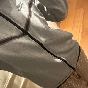 Grå zipjacka från Nike tech med svarta detaljer - Säljer en grå zipjacka från Nike med svarta och svarta detaljer längs bröstet och ärmarna. Jackan har hel dragkedja framtill, lång ärm och en extra ficka med dragkedja på ena ärmen. Perfekt för en sportig och clean look.
