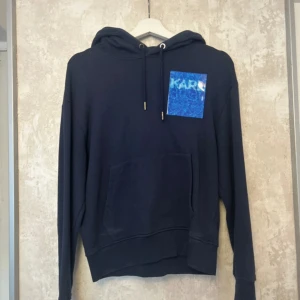 Mörkblå hoodie från Karl Lagerfeld XS - Snygg mörkblå hoodie från Karl Lagerfeld med huva och snörning. Framtill finns en blå patch med logga och en stor magficka. Diskret logotyp även på huvan. Perfekt för dig som gillar stilrena streetwear-plagg.