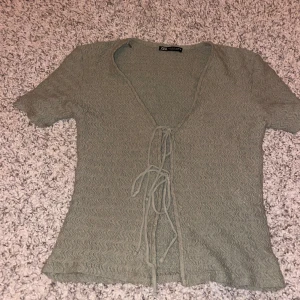 Grön omlottopp med knyt från Zara - Säljer en ljusgrön omlottopp från Zara med korta ärmar och snygg knytning framtill. Toppen har vågig struktur i tyget och djup v-ringning. Perfekt att styla med jeans eller kjol för en trendig look.