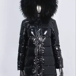 Svart glansig pufferjacka med stor luva - Säljer en svart, glansig pufferjacka med stor fluffig luva i fuskpäls. Jackan är lång, har quiltad design och stängs med dragkedja framtill. Perfekt för kalla dagar när du vill hålla stilen och värmen. Långärmad och riktigt statement med sin oversized luva.