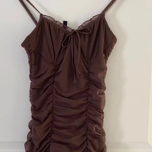 Brun ruched klänning från H&M Divided - Supersnygg brun kort klänning från H&M Divided med smala axelband, rynkade detaljer längs hela klänningen och en söt knytning framtill. Tillverkad i stretchigt syntetmaterial som sitter tight och framhäver figuren.