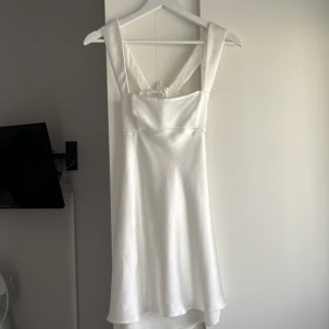 Vit satin klänning med rosett - Superfin vit klänning i satin med breda axelband och snygg rosettdetalj i ryggen. Klänningen har en öppen rygg. Perfekt student eller som vanlig sommarklänning.  Köpte i Spanien på Zara för 400kr och den har aldrig använts, bara provat hemma.