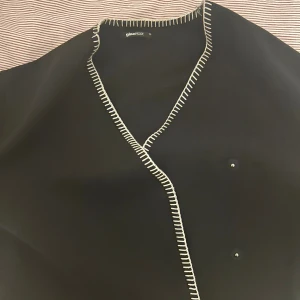 Svart cardigan jacka från Gina Tricot - Svart cardigan jacka från Gina Tricot med vita dekorativa sömmar längs kanterna och ärmsluten. Jackan har v-ringad hals och knappar framtill. Perfekt för dig som gillar stilrena detaljer och vill ha en chill look.