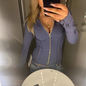 Lululemon Define Jacket - Helt ny Sade/Gold träningsjacka från Lululemon med guldfärgade dragkedjor och snygga sömdetaljer. Jackan har två fickor med dragkedja, lång ärm och hög krage. Perfekt för dig som gillar sportig och clean stil. Kvittot finns och medkommer vid köp! 💕