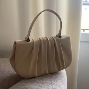 Beige handväska från Perfetto - Snygg beige handväska från Perfetto med mjuk satinfinish och elegant veckad framsida. Väskan har ett stilrent handtag och magnetlås.