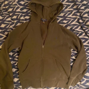 Olivgrön hoodie från Polo Ralph Lauren - Olivgrön hoodie från Polo Ralph Lauren med dragkedja, huva och känguruficka. Klassisk broderad logga på bröstet och mjukt material som känns skönt mot huden. Perfekt för dig som gillar stilrena och bekväma plagg. Hoodien är använd max 10 gånger, nästan nyskick, inget fel på den, storlek L i barn storlek.