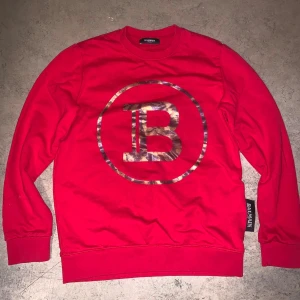Balmain sweatshirt  - Röd sweatshirt från Balmain med stort metallicfärgat B-logga på bröstet. Tröjan har rund hals, långa ärmar och ribbade muddar. Perfekt statement-plagg för dig som gillar exklusiva streetwear-vibbar. Möts i Sthlm