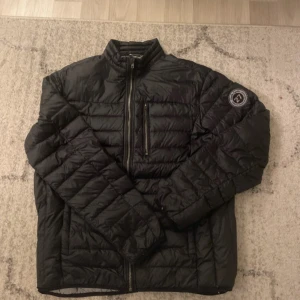 Svart pufferjacka från Hampton Republic - Säljer en svart pufferjacka från Hampton Republic i storlek L. Jackan har quiltad design, hög krage och dragkedja framtill samt en bröstficka med dragkedja. Perfekt för kyliga dagar och enkel att matcha med olika outfits.