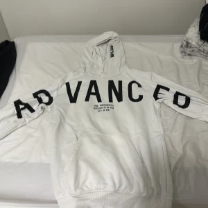 Vit hoodie från Woxo med tryck - Vit hoodie från Woxo med stor svart text 'ADVANCED' över bröstet och ärmarna. Har huva, känguruficka och svart tryckdetalj på luvan. Perfekt för dig som gillar streetwear och vill sticka ut med en clean look.