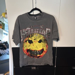 Hellstar t-shirt - Säljer en grå t-shirt från Hellstar i bomull med stort, slitet smileytryck i gult, rött och svart framtill och coolt flames-tryck med stjärna på ryggen. Oversized fit och kortärmad modell, perfekt för dig som gillar streetwear vibes.