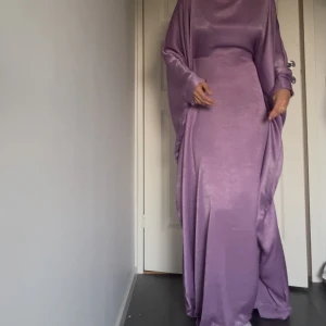 Lila långklänning i satinlook - Säljer en lila långklänning med glansig satinlook och vida, svepande ärmar. Flowig bekväm och elegant klänning, aldrig använd pga färgen faller mig ej i smaken