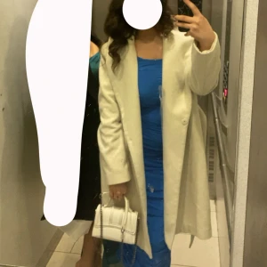 Crémevit kappa med enkel knäppning - Stilren crémevit kappa med rak passform och enkel knäppning framtill. Långärmad modell som går ner över låren, perfekt för att ge en clean och elegant look. Kappan har klassisk krage och är lätt att matcha med olika outfits.