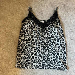 Leopardmönstrad linnetopp med spets - Snygg linnetopp från Samsa Collection i leopardmönster med svarta och beige toner. Toppen har tunna axelband och v-ringning med svart spetskant som ger en extra detalj. Perfekt för dig som vill sticka ut med ett djurmönster och cool vibe.