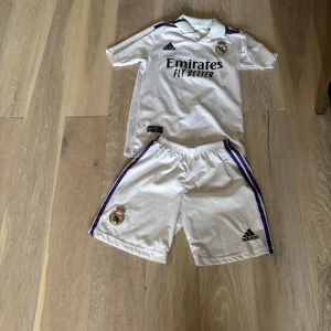 Real Madrid Benzema matchtröja Adidas - Säljer en vit Real Madrid matchtröja med Benzema och nummer 9 på ryggen, tillverkad i ventilerande Climacool-material. Adidas-logga och klubbmärke på bröstet, samt svarta tryck och lila detaljer på axlar och shorts. Setet inkluderar både tröja och shorts.