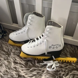 Vita konståkningsskridskor Davos JR 34 - Snygga vita konståkningsskridskor från Davos i storlek 34. Klassisk modell med högt skaft, snörning och svart klack. Yttermaterial i syntet och insida i mjukt tyg. Bladskydd i gult medföljer. Perfekta för dig som vill glida fram på isen med stil.