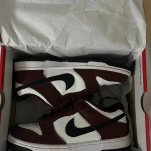 Nike Dunk Low Bordeaux sneakers - Nike Dunk Low Gs sneakers i vinrött och vitt med svart swoosh. Klassisk låg modell med snörning och platt sula. Ovandel i skinn och detaljer i vinröd och vit färg, perfekt för dig som vill sticka ut med stilrena sneakers.