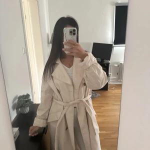 Beige trenchcoat med bälte - Säljer en snygg beige trenchcoat med klassisk krage, knappar framtill och bälte i midjan. Jackan är lång och har stora fickor på sidorna. Perfekt för dig som vill ha en stilren och tidlös look till vardags. Passar till många olika outfits! Köpt på hm, använd i gott skick. Storlek S