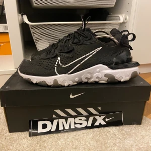 Nike React Vision svart/vit strl 45 - Nike React Vision sneakers i svart med vit sula och stora vita swoosh-detaljer. Ovandelen är i mesh med coola mönster och snörning. Sulan har en unik camo-liknande look och skon är riktigt bekväm för vardagsbruk. Perfekt för dig som gillar streetwear och snygg design.