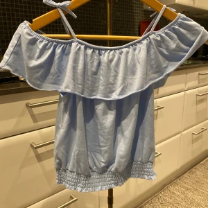 Ljusblå offshoulder topp med volang - Supersöt ljusblå topp med offshoulder och smala axelband som knyts. Volang upptill och smockad nederkant ger en chill och feminin vibe. Perfekt för varma dagar och sommarhäng. Mjuk bomullskänsla och luftig passform.