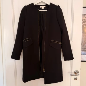 Svart kappa från H&M med dragkedjor - Stilren svart kappa från H&M i storlek 32. Jackan har raka linjer, lång ärm och snygga guldfärgade dragkedjor vid fickorna och framtill. Perfekt för dig som gillar minimalistisk och clean look.