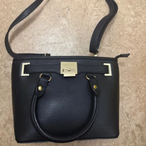 Svart väska från Don Donna - Stilren svart handväska från Don Donna i skinnimitation med både handtag och axelrem. Väskan har guldfärgade metalldetaljer. Aldrig använt och så gott som ny, pris går att diskutera. 