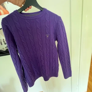 Lila kabelstickad tröja från Gant - Säljer en lila kabelstickad tröja från Gant med rund halsringning och broderad logga på bröstet. Tröjan har lång ärm och ribbade muddar, perfekt för dig som vill ha en klassisk och färgstark look. Står att den är M men passade inte mig så den är mer åt S hållet.