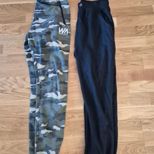 Camo och svarta mjukisbyxor - Två par mjukisbyxor, ett par med grön camouflage-mönster och dragkedja vid fickan samt ett par helsvarta. Båda har resår i midjan och är perfekta för chill eller träning. Camobyxan har vit logga på låret.