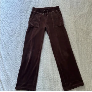 Bruna byxor Juicy Couture XXS - Mysiga bruna velourbyxor från Juicy Couture med broderad logga på fickan, snörning i midjan och fickor både fram och bak. Byxorna har en rak passform. Byxorna är uppsydda så de är kortare än vanligt.💘De är ungefär 65cm från grenen till slutet av benet.