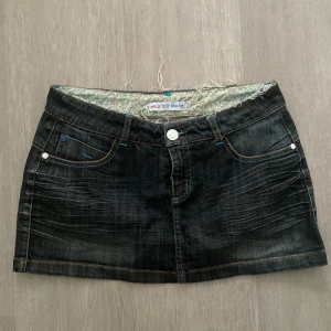 Mörk jeanskjol från Wild Jeans Italy - Snygg mörkblå jeanskjol med slitningar och kontrastsömmar i orange och blått. Kjolen har klassisk femficksdesign, bälteshällor och coola dragkedjor på bakfickorna. Perfekt för dig som gillar en edgy och trendig look.