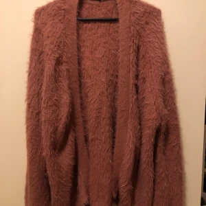 Luddig rosa kofta med lång ärm - Supermjuk och fluffig rosa kofta med lång ärm. Koftan är öppen framtill och har en mysig, lurvig textur som gör den perfekt att slänga på sig när det är kyligt. Passar dig som gillar en chill och avslappnad stil.