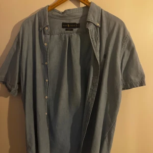 Blå kortärmad skjorta Ralph Lauren XL (slim fit) - Snygg ljusblå kortärmad skjorta från Ralph Lauren i slim fit. Skjortan har klassisk krage, knappar framtill och diskret broderad logga på bröstet. Tillverkad i mjuk bomull. Present som inte passar aldrig använd