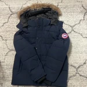 Mörkblå dunjacka från Canada Goose - Säljer en mörkblå dunjacka från Canada Goose med avtagbar huva och fuskpäls. Jackan har klassisk patch på ärmen, dragkedja och knappar framtill samt ribbade muddar. Perfekt för kalla vinterdagar och riktigt snygg passform.