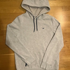 Ljusgrå hoodie från Hollister - Snygg ljusgrå hoodie från Hollister med diskret logga på bröstet. Tröjan har huva med svarta snören, magficka och långa ärmar med ribbade muddar. Perfekt för chill dagar och enkel att matcha med jeans eller joggers.