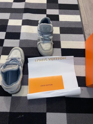 Louis Vuttion skor helt nya - Säljer ett par Louis Vuitton sneakers i ljusgrått och vitt med klassiskt LV-monogram i tyget. Skorna har snörning, rund tå och detaljer i mocka och skinn. Snygg kontrast mellan olika material och tydlig LV-logga på plös och häl. Perfekta för dig som gillar exklusiva streetwear-vibbar.