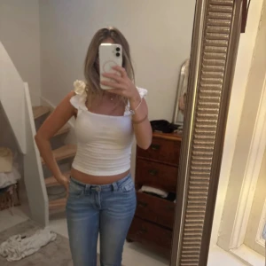 Vit croppad linnetopp med volang - Säljer en vit croppad linnetopp med volangdetaljer på axelbanden. Toppen har fyrkantig halsringning och är figurnära med ribbad struktur. Perfekt för dig som gillar en söt och fräsch stil till sommaren.