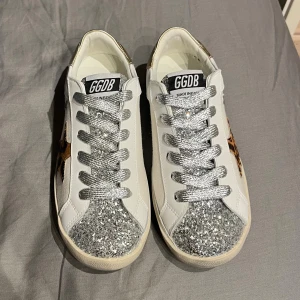 Golden Goose sneakers med glitter och leopard - Golden Goose sneakers i vitt skinn med silverglittrande snörning och tå, guldiga detaljer på hälen och svart glitterrand bak. Sidan har en leopardmönstrad stjärna som ger extra edge. Platt sula och rund tå, perfekt för dig som gillar att sticka ut.