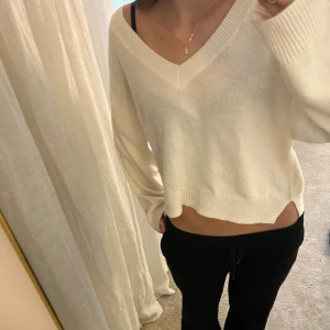 Vit croppad stickad tröja med v-ringning - Säljer en vit croppad stickad tröja med bred v-ringning och långa ärmar. Tröjan har ribbade muddar vid hals, ärmslut och nederkant, vilket ger en chill och avslappnad vibe. Perfekt för dig som gillar en loose passform och vill ha något mjukt och bekvämt.