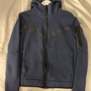 Blå hoodie från Nike med dragkedja - Blå hoodie från Nike med hel dragkedja framtill och huva. Mjuk och skön, perfekt för chill eller träning. Klassisk Nike-logga på bröstet.