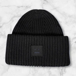 Svart ribbad mössa från Acne Studios - Svart stickad mössa med bred uppvikt kant och Acne Studios klassiska ansiktslogga framtill. Ribbstickad design som ger en snygg och chunky look, perfekt för kalla dagar. Materialet är mjukt och värmande, och färgen är helsvart.