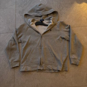 Grå hoodie från Polo Ralph Lauren zip - Grå hoodie från Polo Ralph Lauren med dragkedja och huva. Insidan är mjuk och fleecefodrad, perfekt för kyliga dagar. Klassisk och enkel design med långärmade ärmar. Size S och kvalité 7/10. Skriv om ni har några frågor