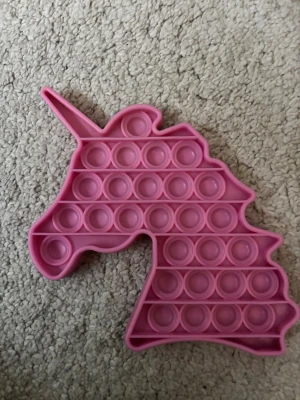 Unicorn Pop It Fidget Toy - Rosa Pop It fidget-leksak i form av en enhörning. Tillverkad i mjuk silikon, perfekt för stresslindring och sensorisk lek. Produkten är i gott skick med endast mindre tecken på användning. Rolig och tålig för både barn och vuxna.